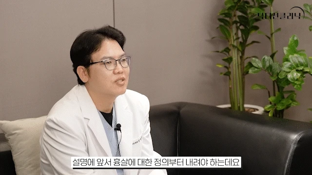 흰색 가운을 입은 남성 의사가 안경을 쓰고 마이크를 착용한 채 소파에 앉아 비절개흉살치료레이저와 흉살의 정의에 대해 말하고 있습니다. 배경에 식물이 보입니다.